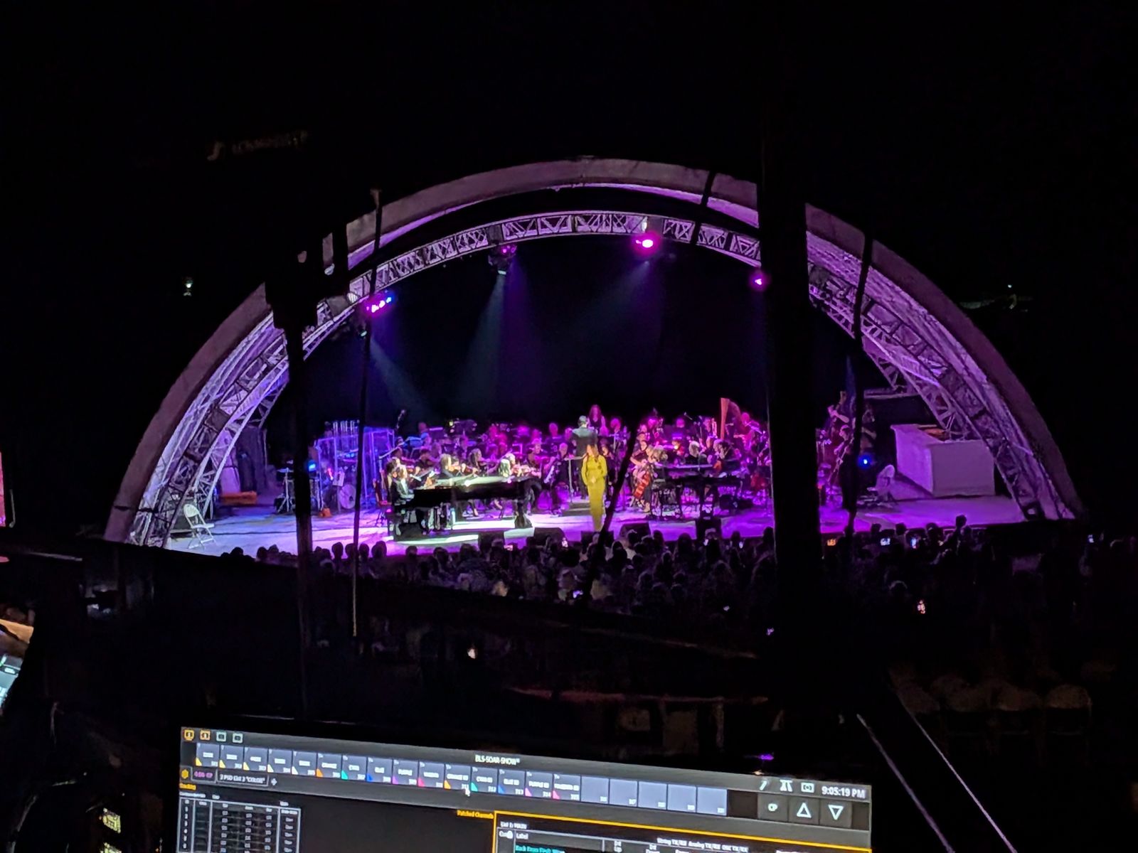 Outdoor orchestra concert under an arched truss shell, lighting console in the foreground